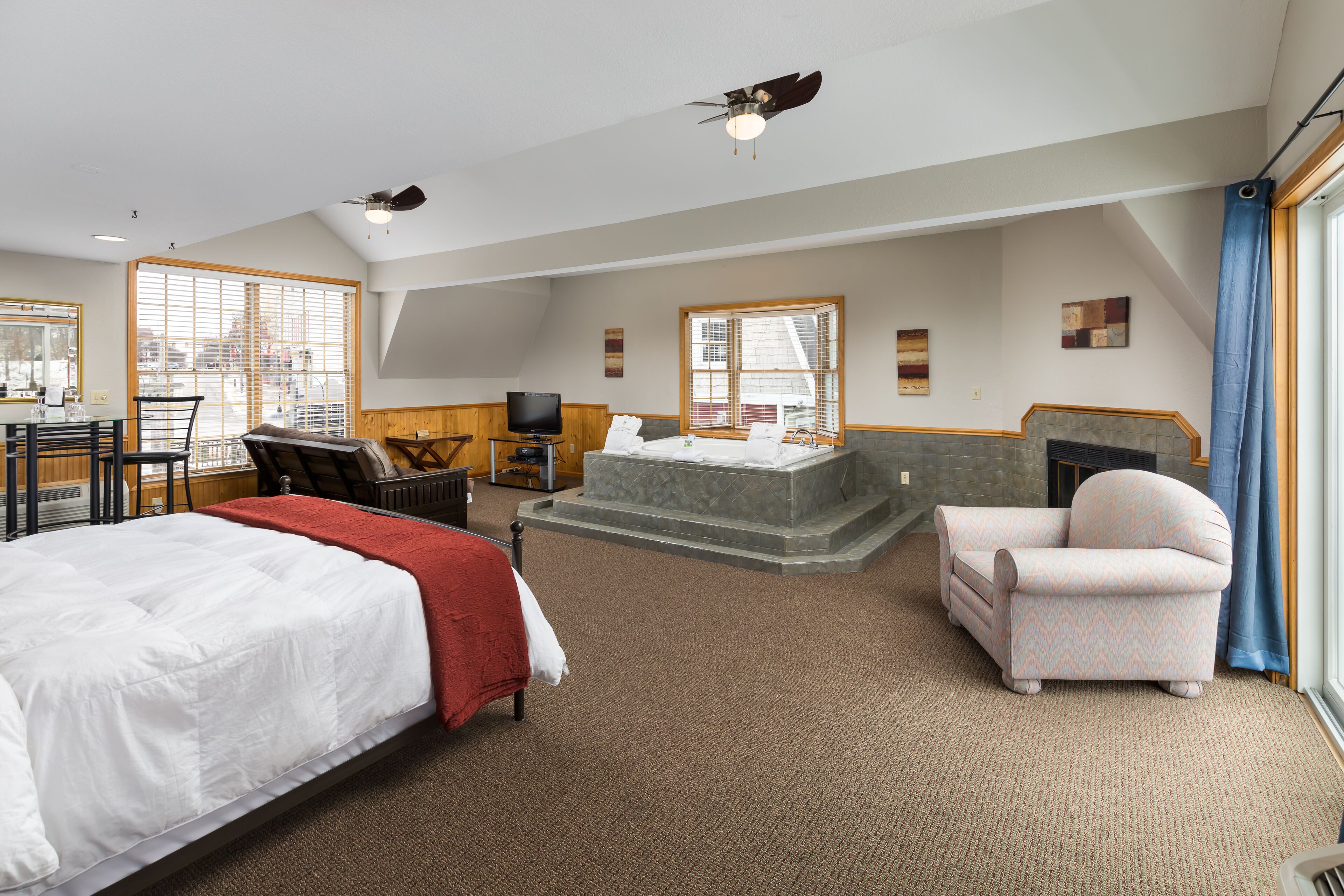 A spacious hotel room with a large bed, a seating area with a chair and a small table, a fireplace, and a large window providing a view of the city.