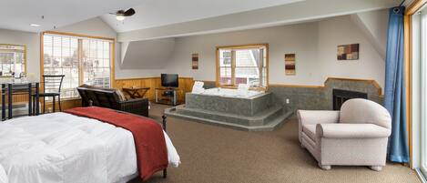 A spacious hotel room with a large bed, a seating area with a chair and a small table, a fireplace, and a large window providing a view of the city.
