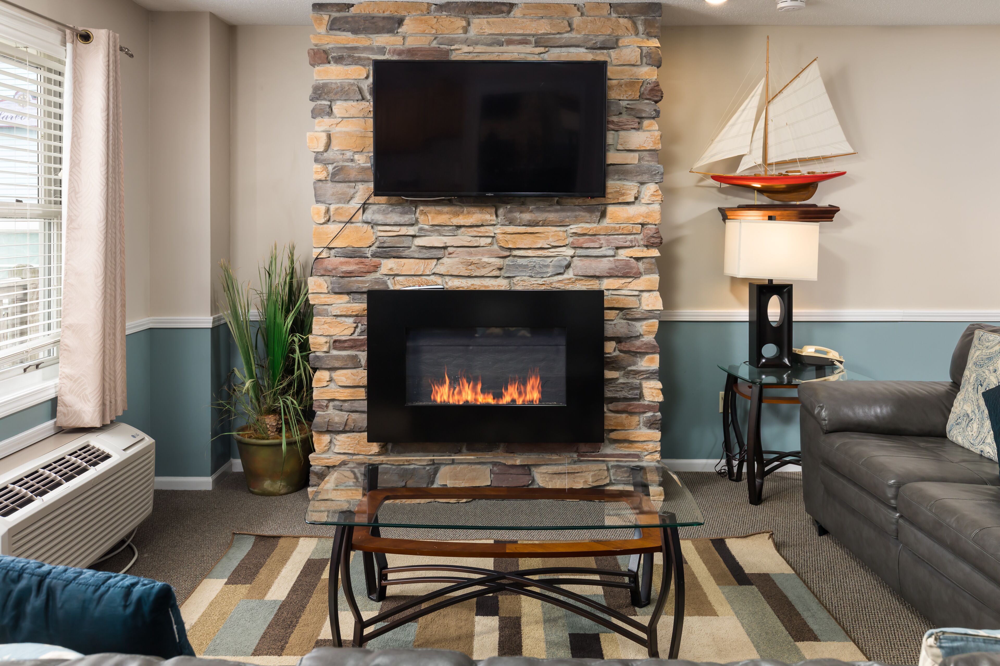 A living room with a stone fireplace, a flat-screen TV above it, and a sailboat decoration on the wall.