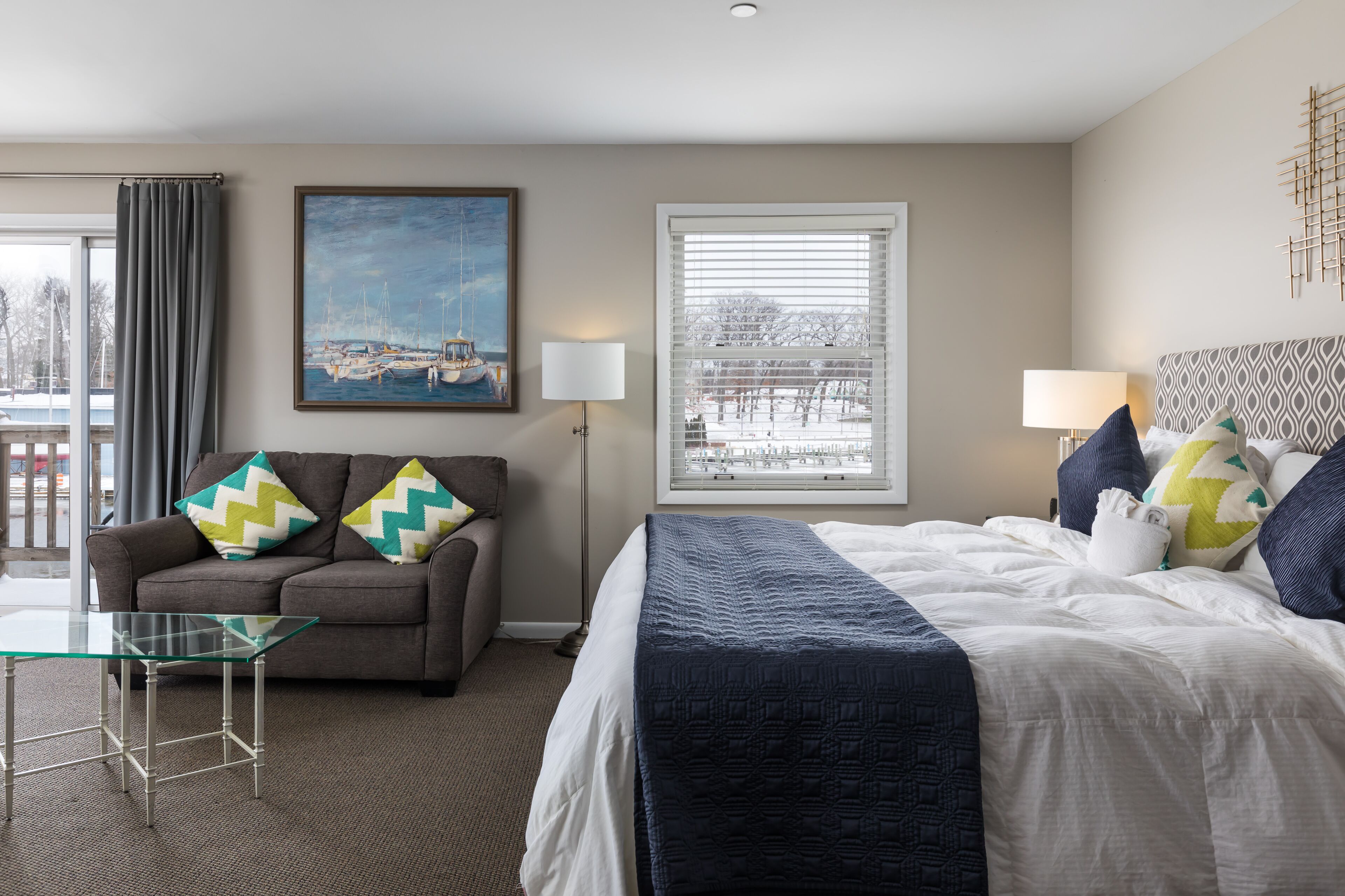 A hotel room with a bed, a sofa, a glass coffee table, a painting, a window with blinds, and a lamp.