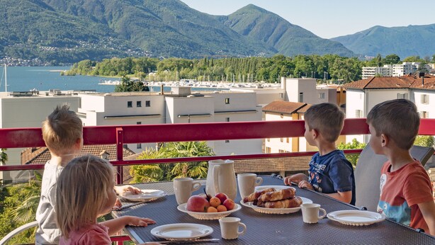 A terrace with a table set for breakfast, overlooking a mountainous landscape and a body of water. - APART HOLIDAYS Residenza Flora (Muralto)