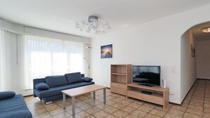 A living room with a sofa, a coffee table, a TV on a wooden stand, and a chandelier. - APART HOLIDAYS Residenza Flora (Muralto)