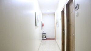 A hallway with white walls, a tiled floor, and a fire extinguisher mounted on the wall. There is a door with a number sign on it. - Hotel Grandeur (Mumbai)