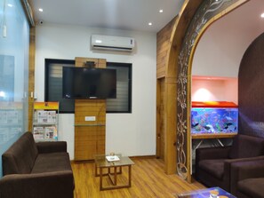 A hotel room with a wooden floor, a flat-screen TV mounted on a wooden stand, a glass coffee table, two brown sofas, and a fish tank with colorful fish.
