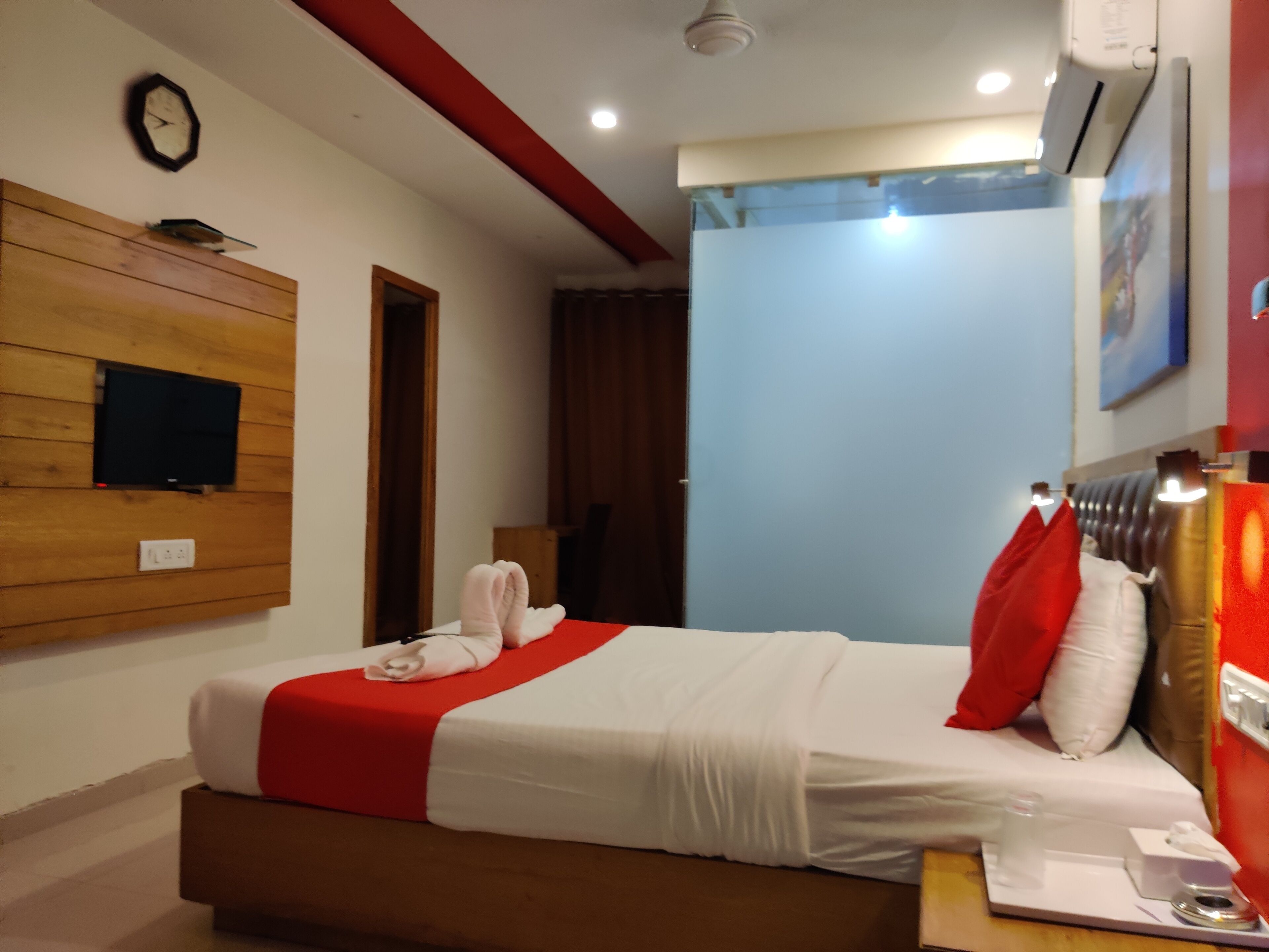 A hotel room with a bed, a flat-screen TV mounted on a wooden panel, a clock on the wall, and a window with a curtain.