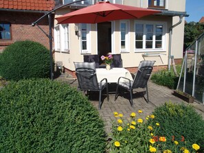 A patio with a table and chairs, a red umbrella, and a flower bed with yellow flowers.