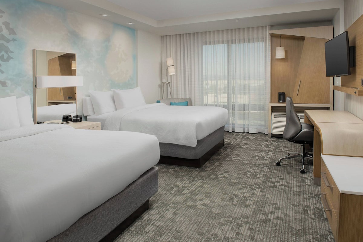 A hotel room with two beds, a desk, a chair, a TV, and a large window with curtains.