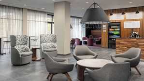 A modern lobby with grey upholstered chairs, a round table, a coffee station, and a menu board. - Courtyard by Marriott Portland East (Portland)