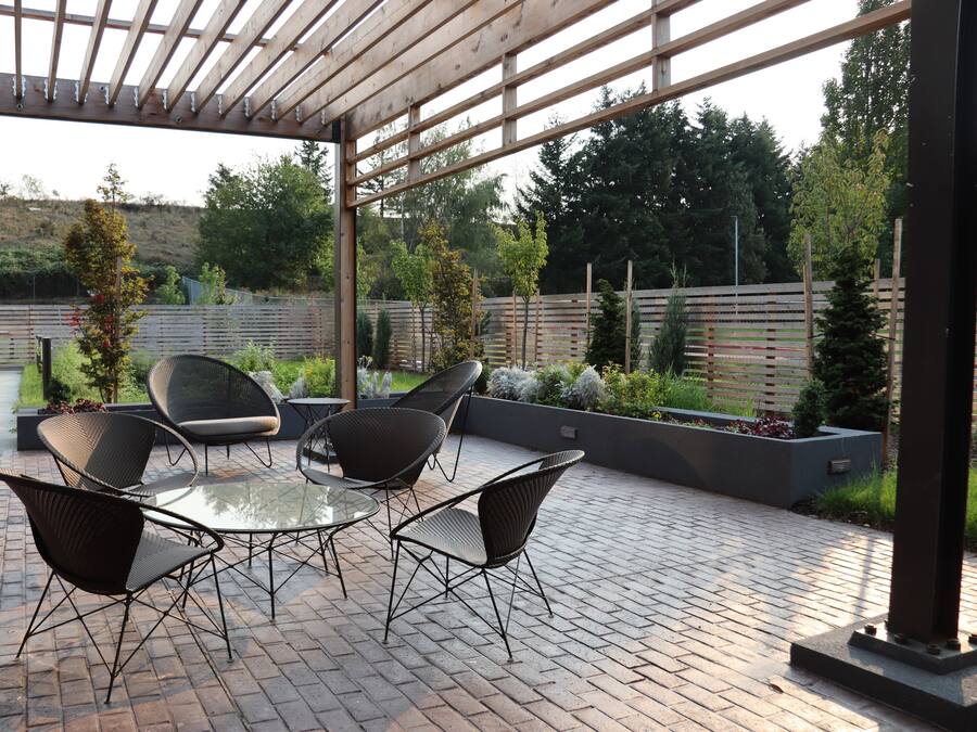 Un patio al aire libre con una mesa de vidrio rodeada de sillas de mimbre, una pérgola en la parte superior y vistas a un jardín ajardinado con valla de madera y árboles.