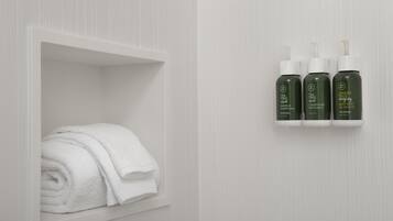 Ein Badezimmer mit einem weißen Handtuch in einer Ablage und drei Flaschen mit Teebaumöl, Zitrone-Salbei und Duschgel an der Wand.