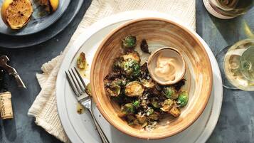 A bowl of roasted Brussels sprouts with a creamy sauce, a fork, and a wine glass on a table.