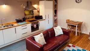 A compact kitchen with a stove, microwave, and sink, a red leather sofa with floral cushions, and a wooden dining table with chairs.