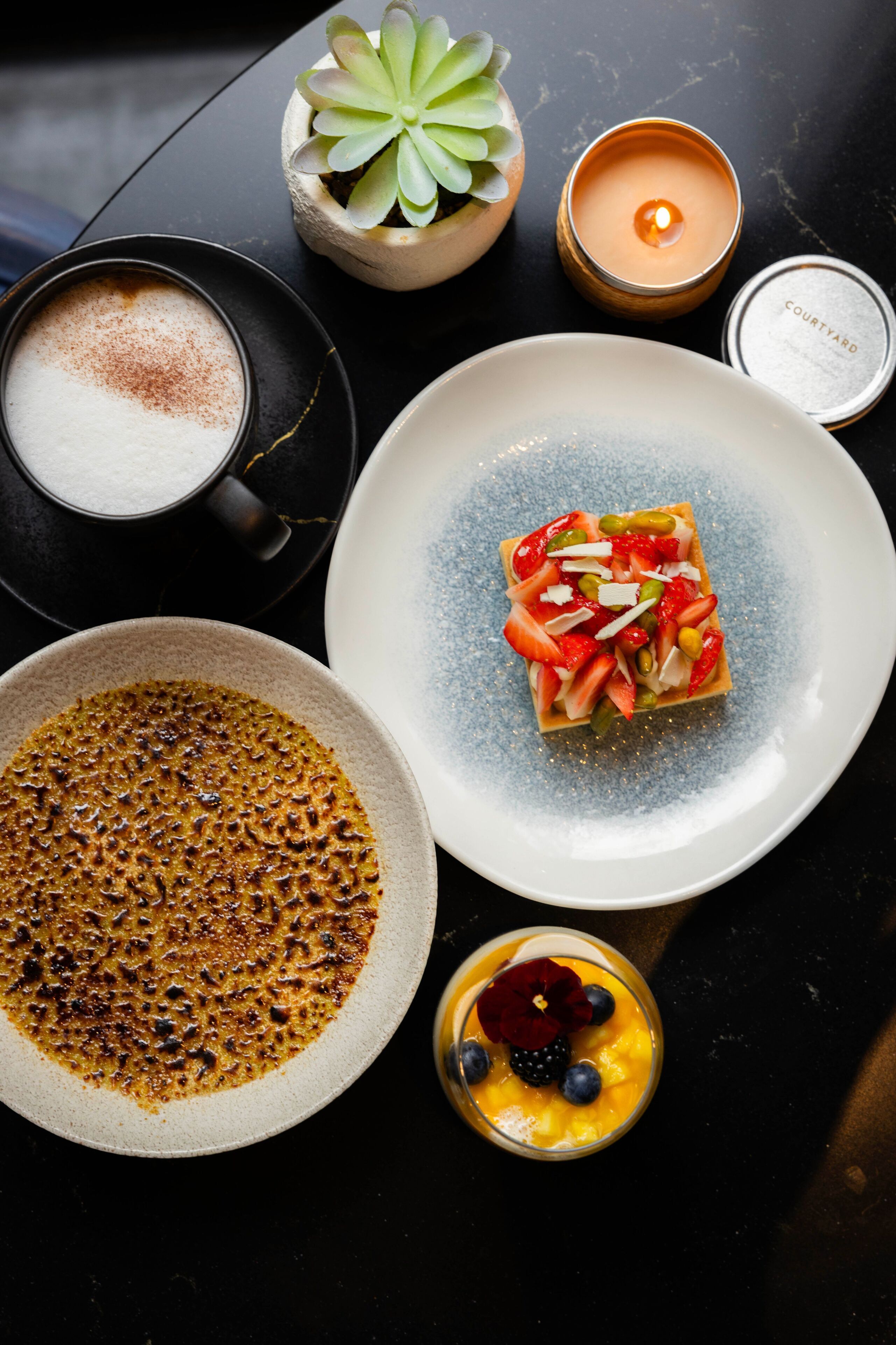 A table with a dessert, a candle, a small plant, a coffee cup, and a bowl of food.