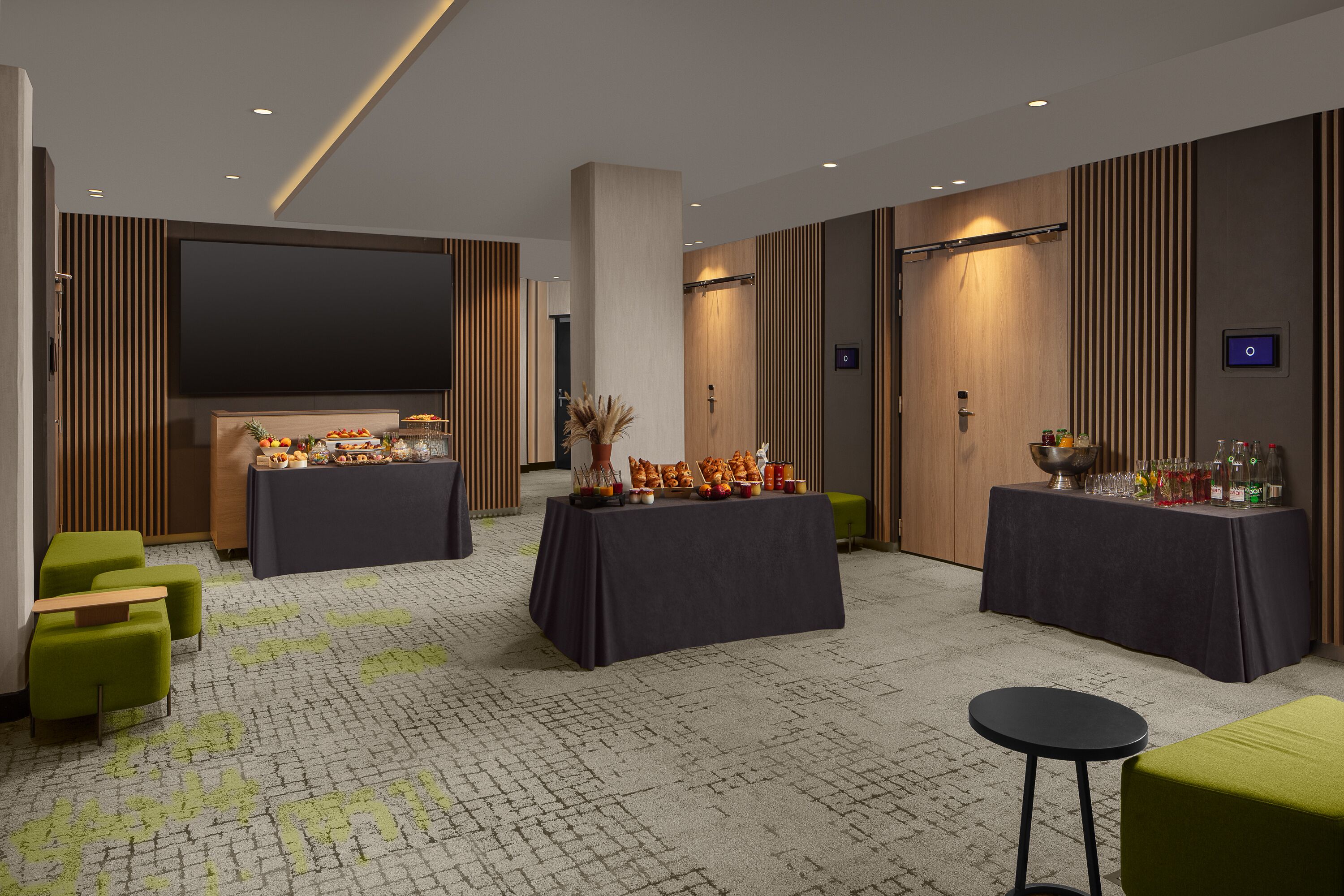 A modern hotel lobby with two round tables set up for dining, green upholstered seating, a large flat-screen TV, and a patterned carpet.