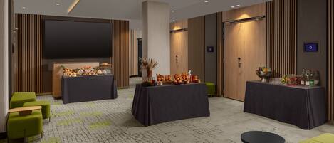 A modern hotel lobby with two round tables set up for dining, green upholstered seating, a large flat-screen TV, and a patterned carpet.