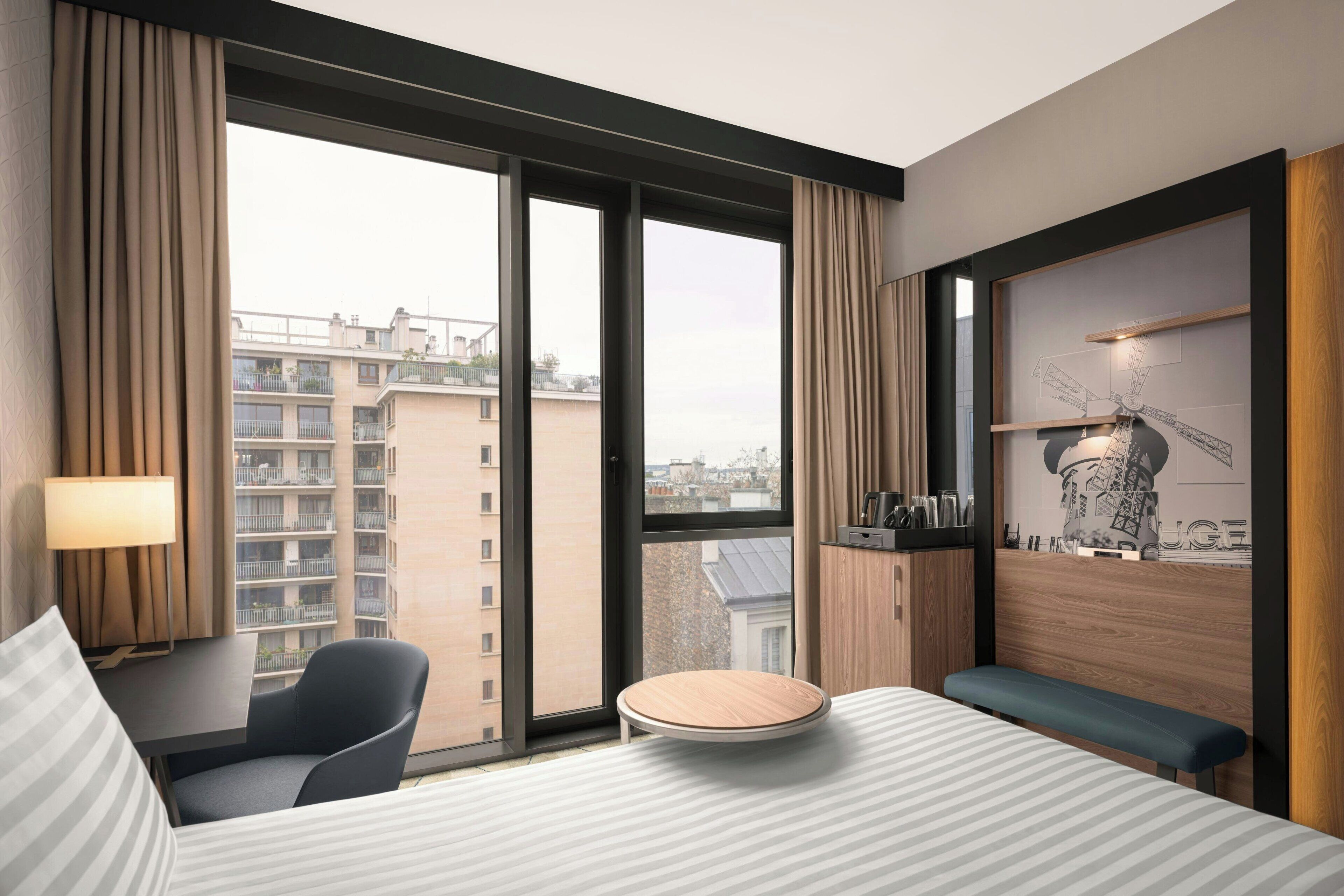 A hotel room with a large window, a round table, a bed, a chair, a lamp, and a wall-mounted shelf with decorative items.