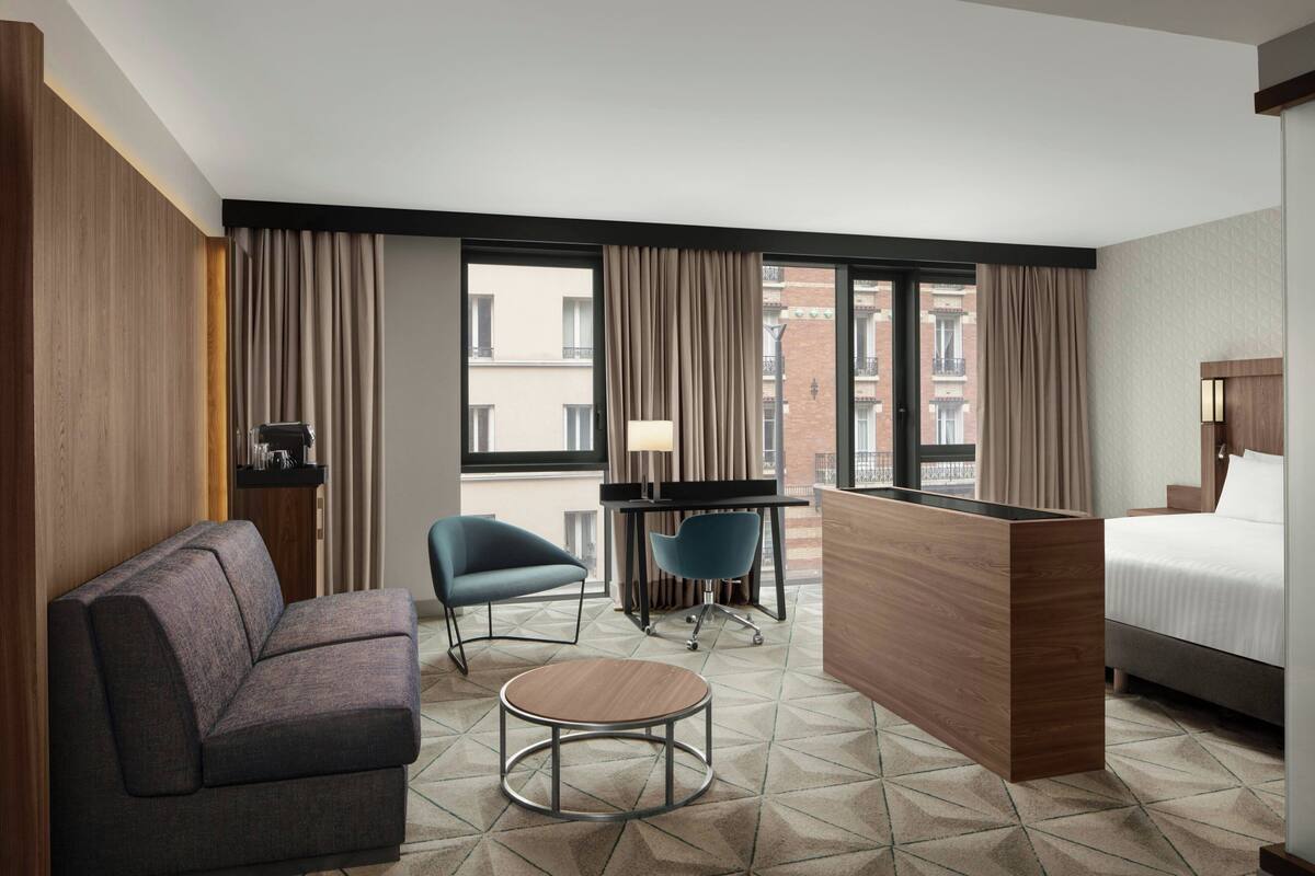 Hotelkamer met een bank, een klein rond tafeltje, een bureau met stoel, een bed en grote ramen met gordijnen.