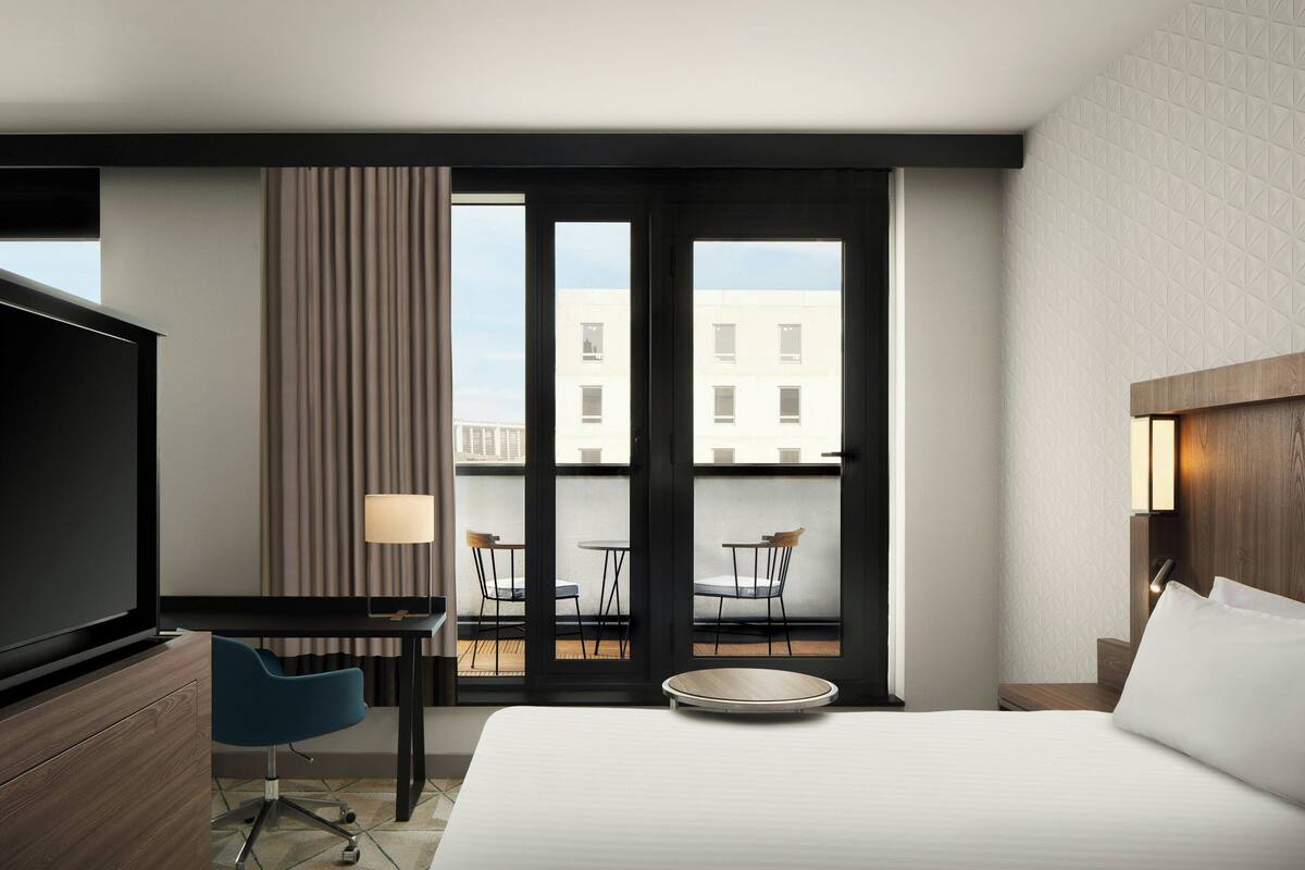 Hotelkamer met een bed, een bureau, een stoel, een televisie en een balkon met uitzicht op gebouwen.