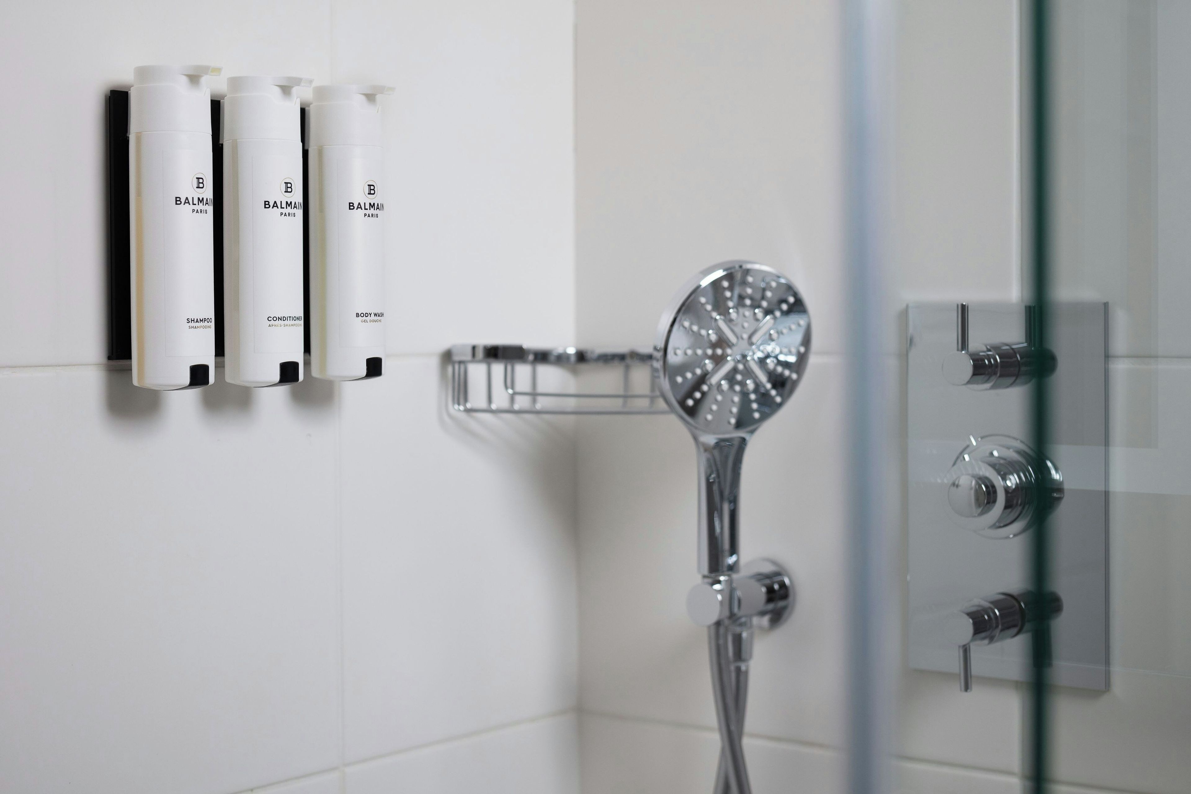 A bathroom with a glass shower enclosure, a handheld showerhead, and three bottles of Balmain Paris shampoo, conditioner, and body wash.