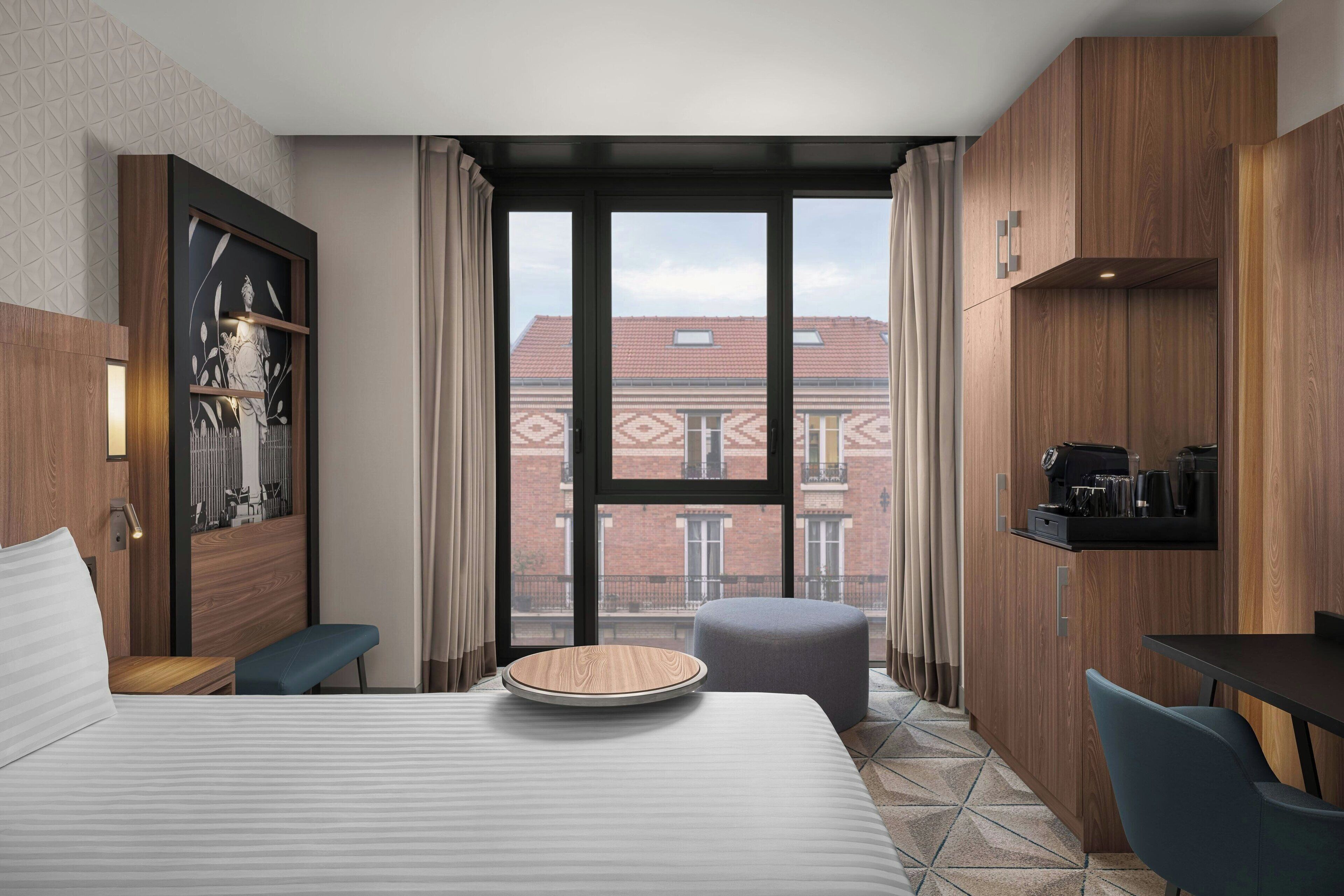 Una habitación de hotel moderna con una cama grande, un escritorio pequeño, una silla, una mesa redonda de madera y vistas a edificios a través de amplios ventanales.