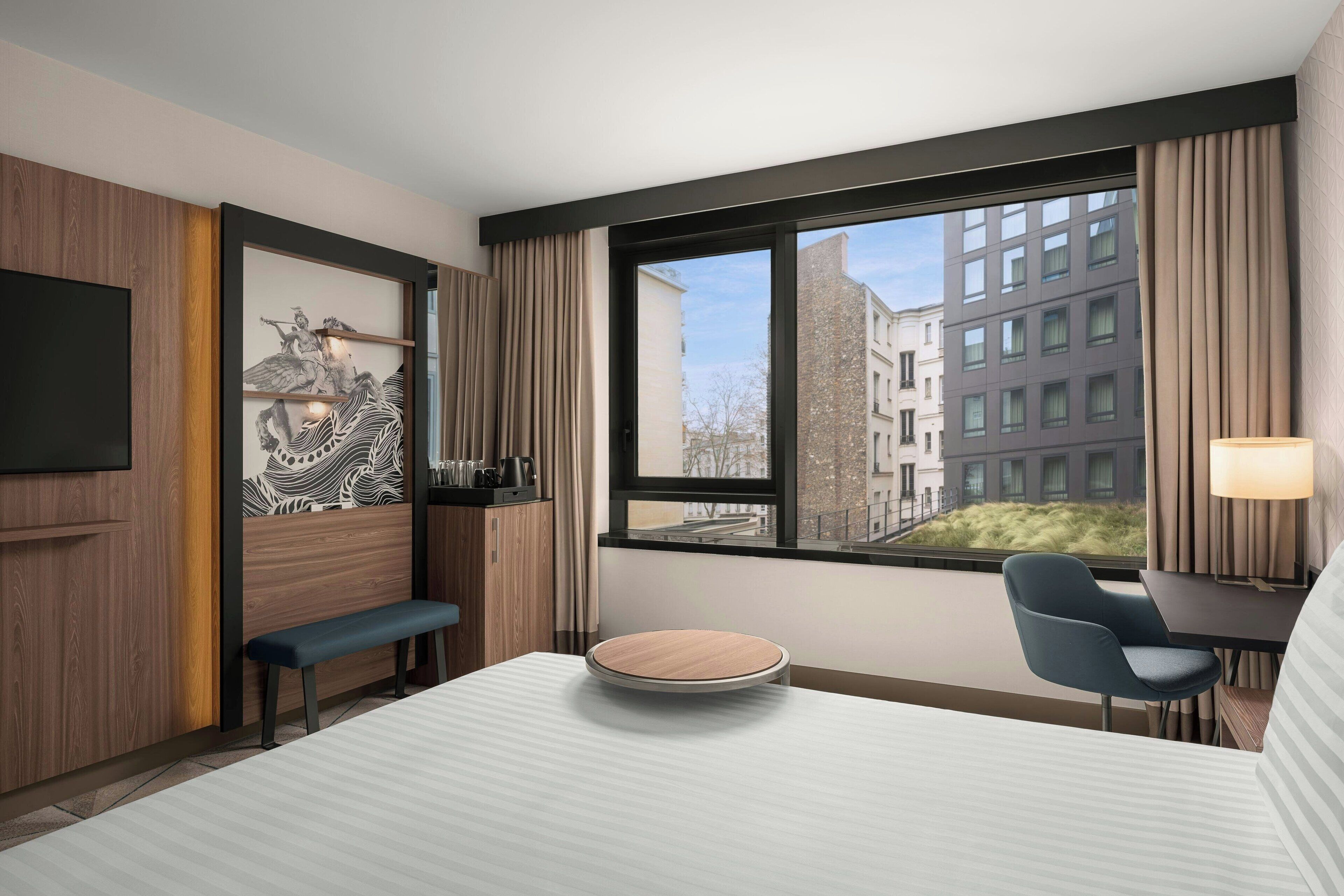 A modern hotel room with a large bed, a desk, a chair, a bench, a TV, and a window with a city view.