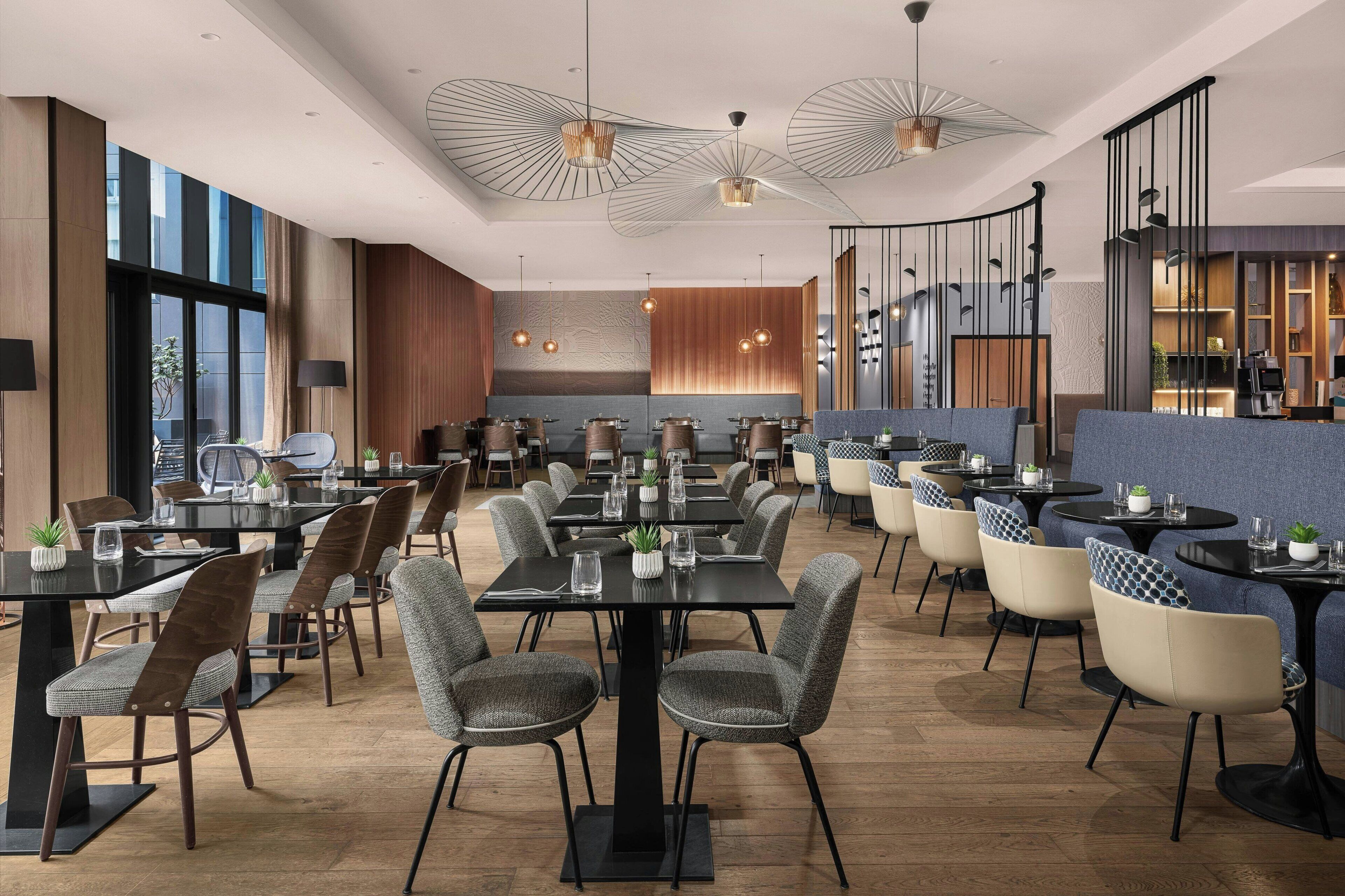 A modern restaurant with multiple dining tables and chairs, large windows, and contemporary lighting fixtures.