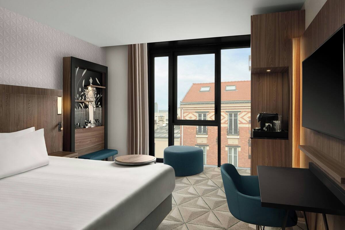 Een moderne hotelkamer met een groot bed, een bureau, een stoel en uitzicht op de stad door grote ramen.