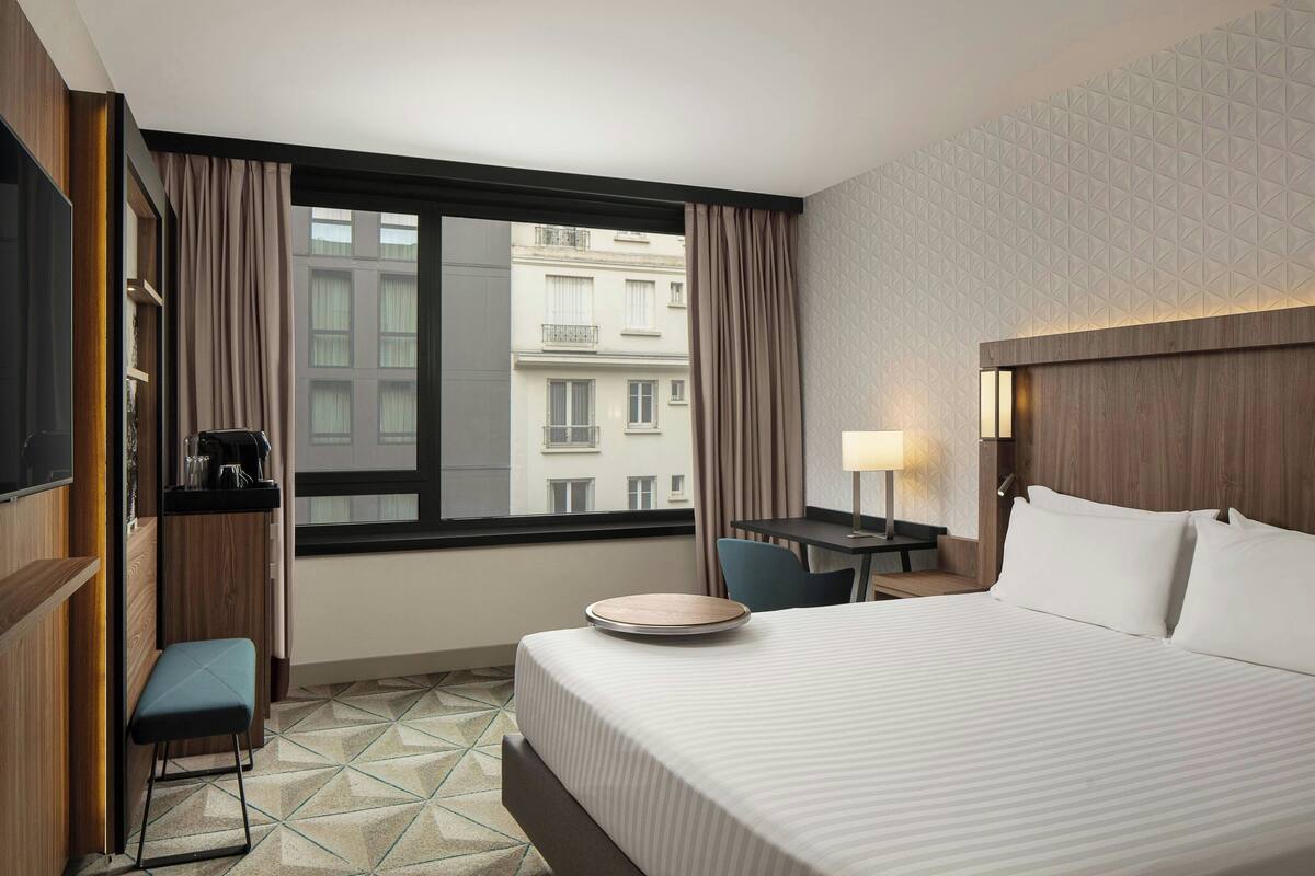 Hotelkamer met een bed, bureau, stoel, een klein krukje, televisie en raam met gordijnen.