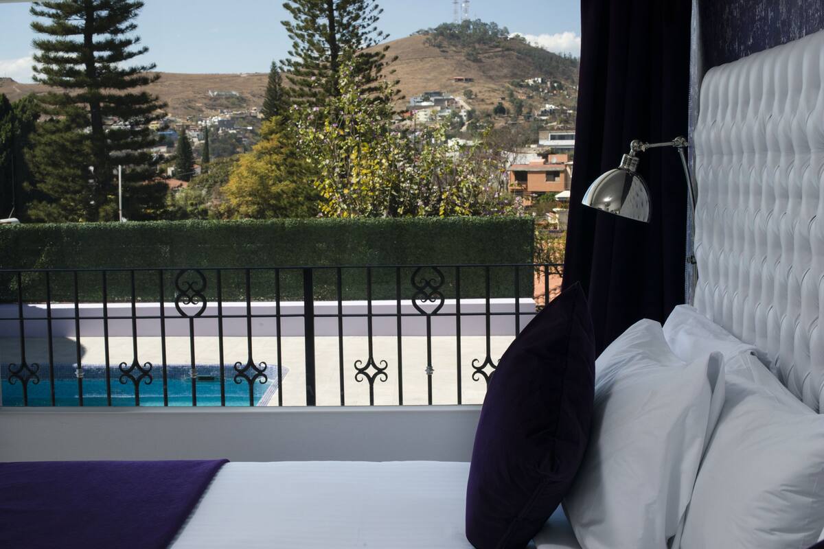 Habitación de hotel con vistas a una piscina, un balcón con baranda metálica y una cama con ropa blanca y una almohada morada.
