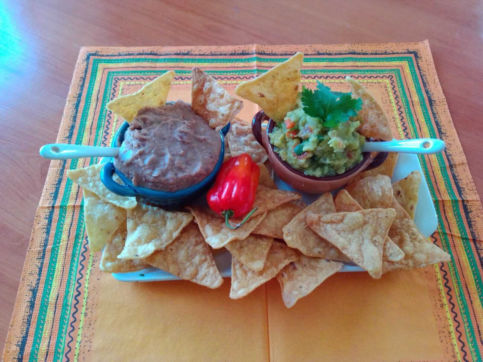 A plate of tortilla chips with two dips, one of black bean and one of guacamole, and a red chili pepper.