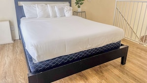 A neatly made bed with white linens and a dark headboard, a small side table with a plant, and a modern abstract painting on the wall.