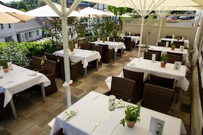 Eine Terrasse im Freien mit Tischen und Stühlen, beschattet durch Sonnenschirme, umgeben von Grünpflanzen.