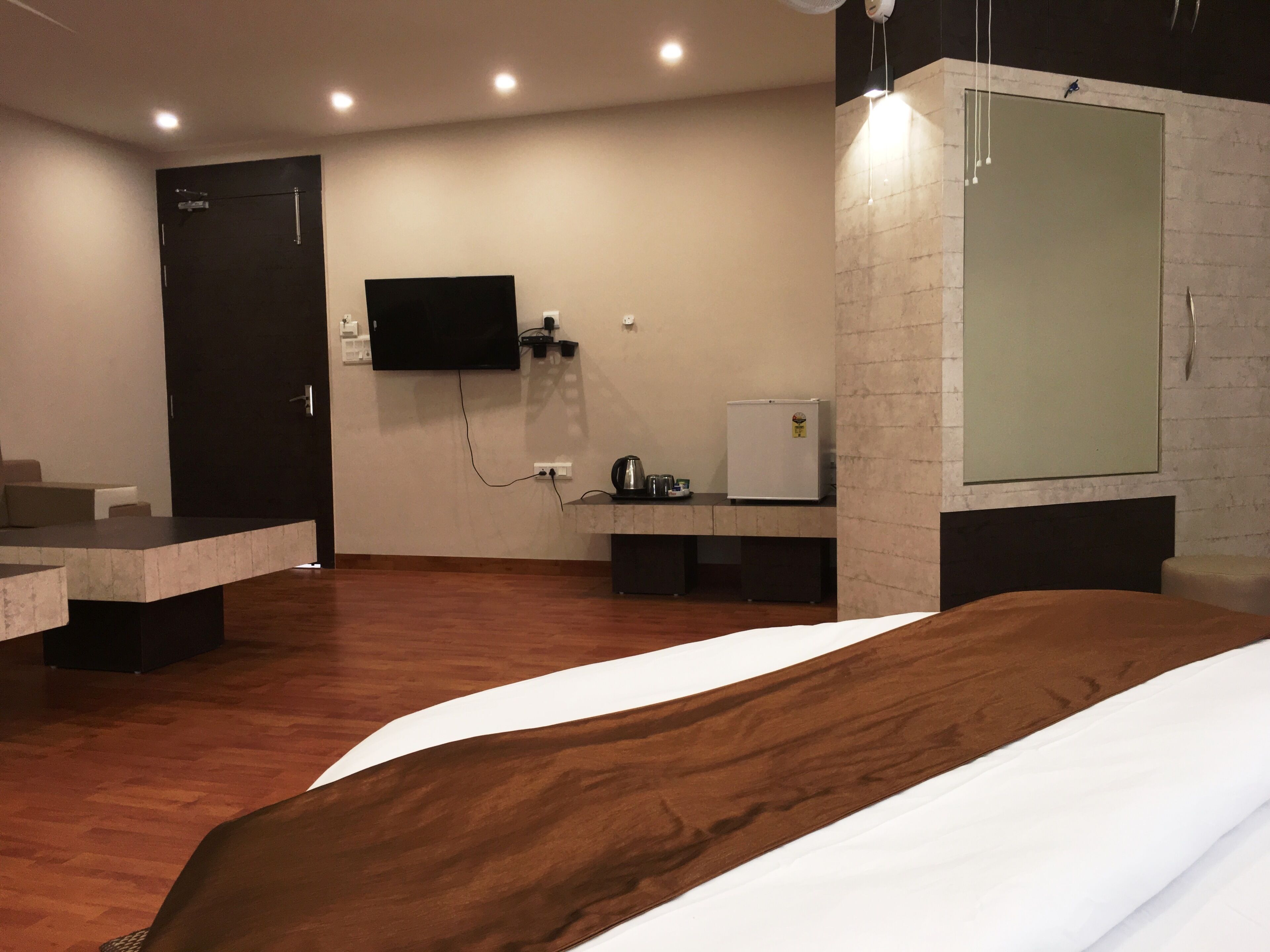 A hotel room with a bed, a TV mounted on the wall, a small refrigerator, and a wooden floor.