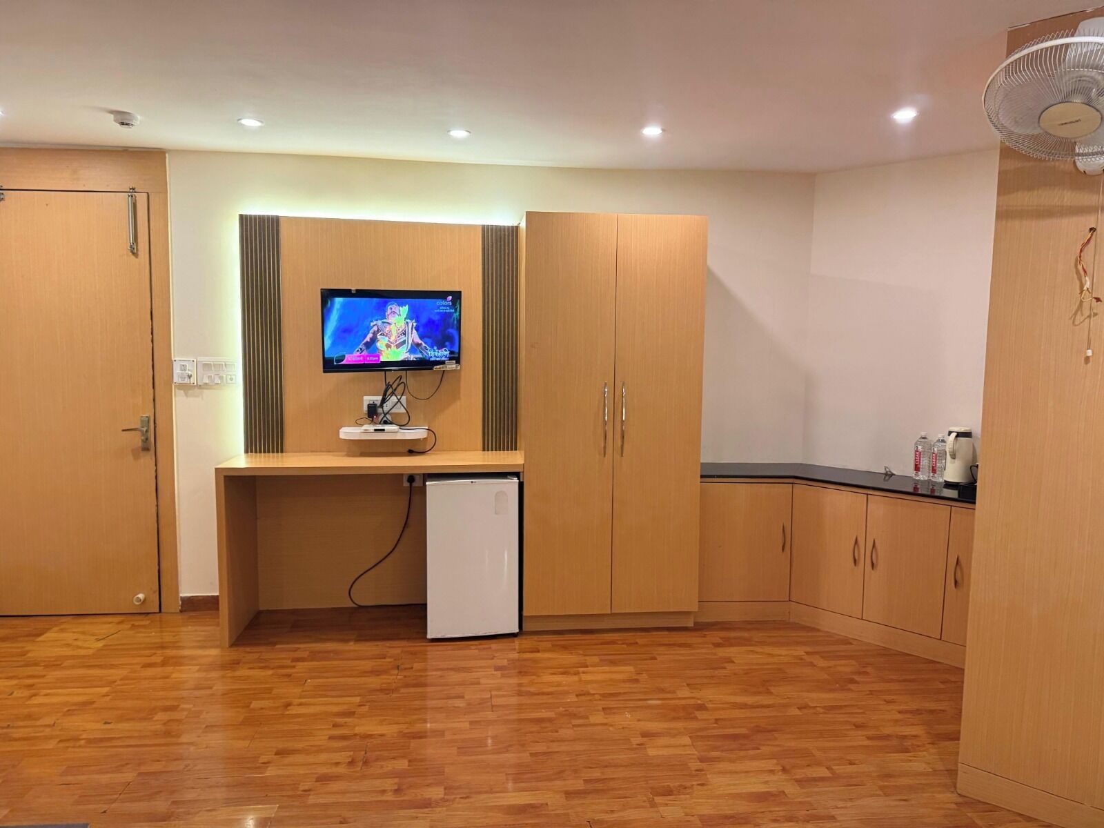A compact room with a wooden floor, a small white refrigerator, a TV mounted on the wall, and wooden cabinets.