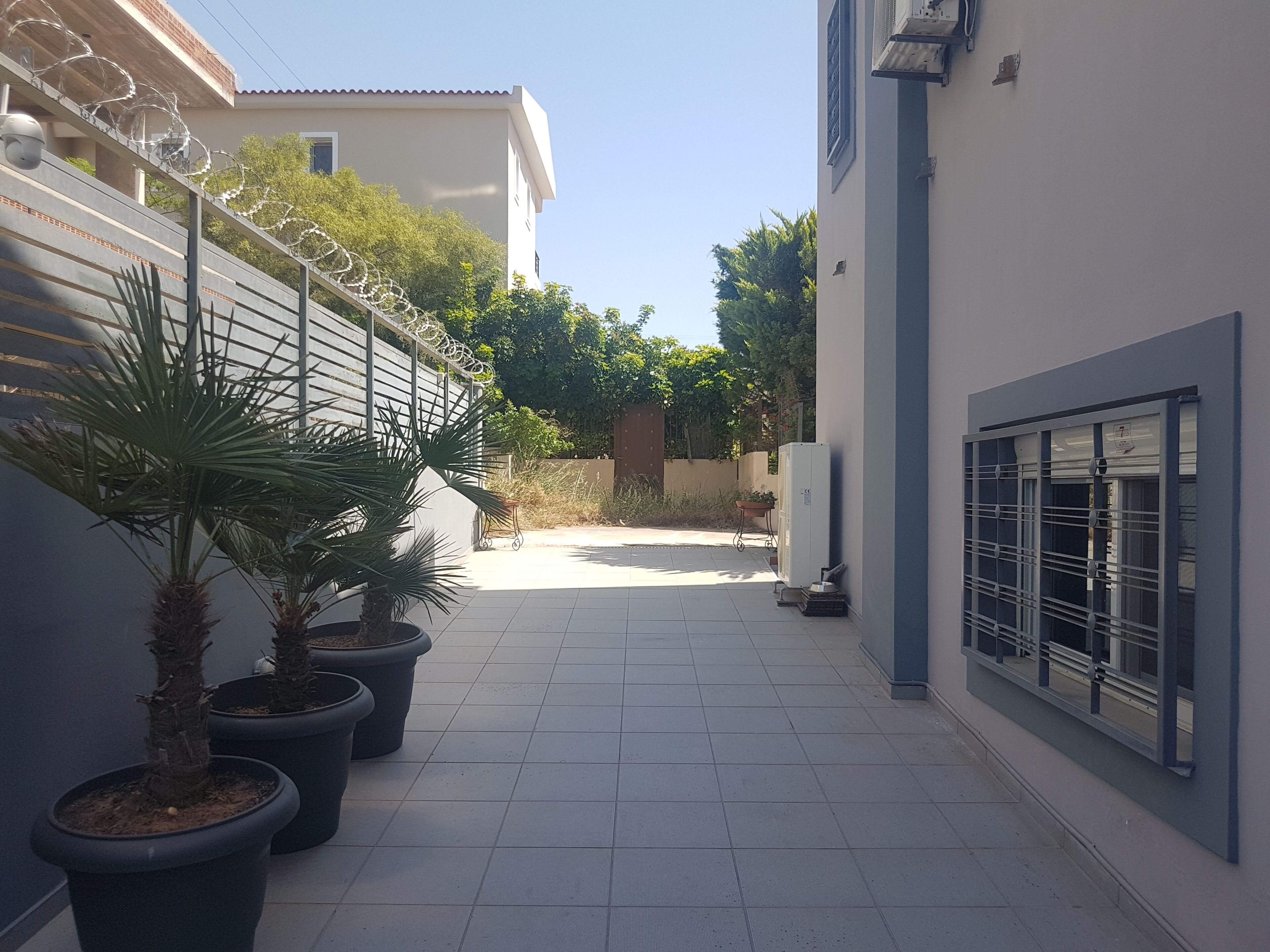 A narrow outdoor corridor with potted plants, a metal fence, and a building with a barred window.
