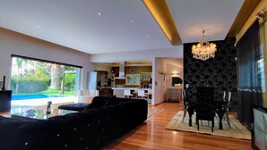 Un salon moderne avec un grand canapé noir, un coin repas agrémenté d’un lustre, et une cuisine en arrière-plan.