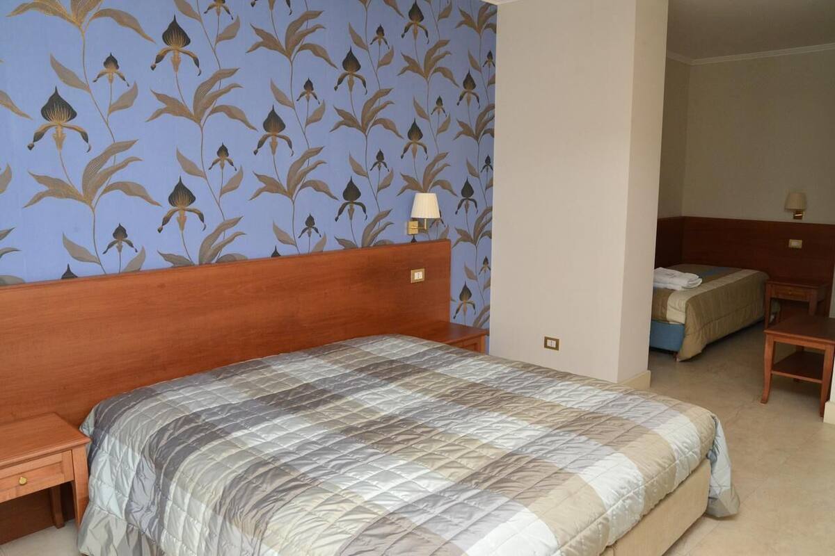 A hotel room with a large bed, a smaller bed, a nightstand, a lamp, and a floral wallpaper pattern.
