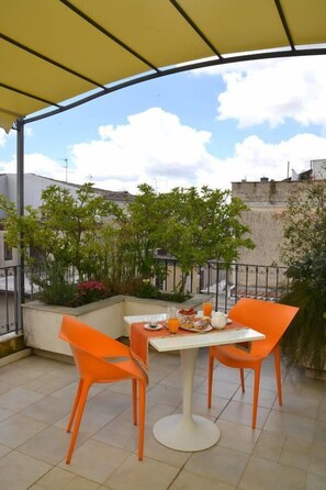 A covered outdoor seating area with a table and two orange chairs, a small table with a bottle and a cup, and a view of a cityscape with buildings and greenery. - B&B Talìa (Grammichele)