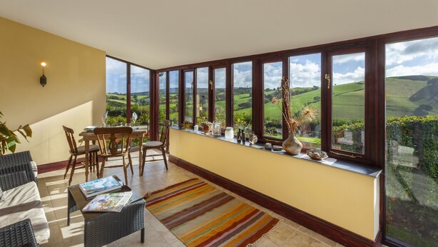 Une pièce spacieuse dotée d’une grande fenêtre donnant sur un paysage magnifique, d’un coin salon confortable avec des meubles en osier et d’une petite table sur laquelle est posé un magazine.