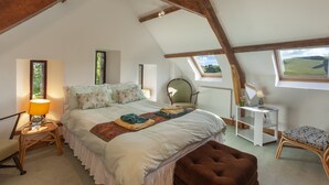 Une chambre avec un plafond en pente, un lit avec une couverture, des oreillers, une lampe de chevet, une chaise, une petite table avec une lampe et une fenêtre donnant sur un beau paysage.