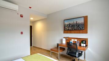 A hotel room with a wooden desk, a chair, a cityscape painting, and a bed with a green mat.