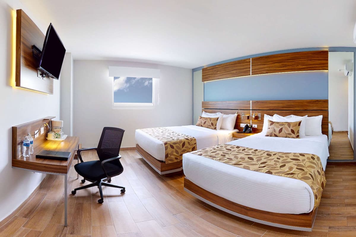 A hotel room with two beds, a desk with a chair, a flat-screen TV mounted on the wall, and a window with a view of the sky.