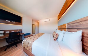 A modern hotel room with a wooden headboard, a large bed with patterned bedding, a desk with a computer, and a flat-screen TV mounted on the wall. - Sleep Inn Queretaro (Querétaro)