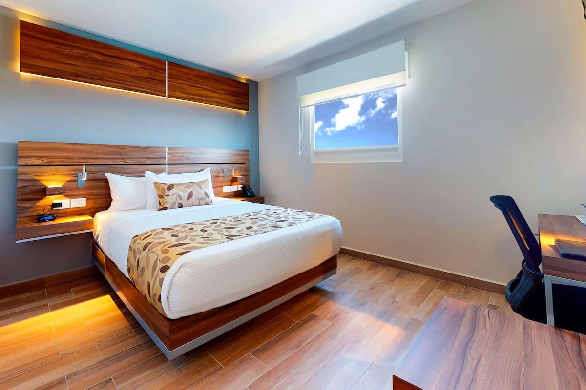 A modern bedroom with a wooden headboard, a large bed with patterned bedding, bedside lamps, and a view of the sky through a window.