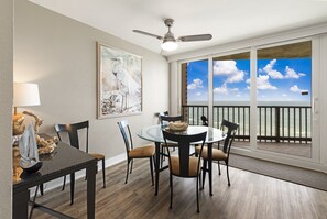 A dining area with a round glass table, four chairs, a ceiling fan, and a large window with a view of the sky and a balcony.