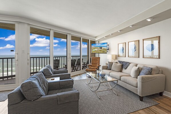 A spacious living area with a large sliding glass door leading to a balcony, a comfortable sofa, a coffee table, and a television mounted on the wall.