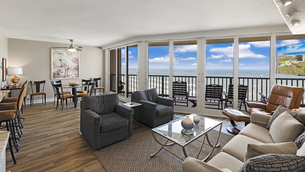 A spacious living area with a balcony overlooking the ocean, furnished with sofas, chairs, and a dining table.
