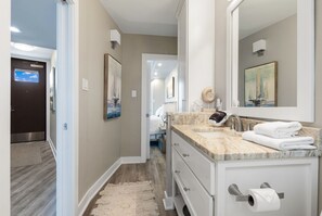 A modern bathroom with a marble countertop, a large mirror, and a view of the bedroom through an open door.