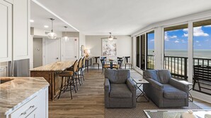 A modern kitchen with a marble countertop, wooden bar stools, and a dining area with a view of the ocean.