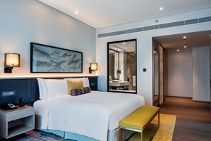 Ein modernes Hotelzimmer mit einem großen Bett, Nachttischlampen, einer Bank und einem durch eine Glastür einsehbaren Badezimmer.